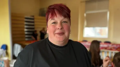 A woman with short red hair standing in a yellow classroom. Students can be seen behind her.