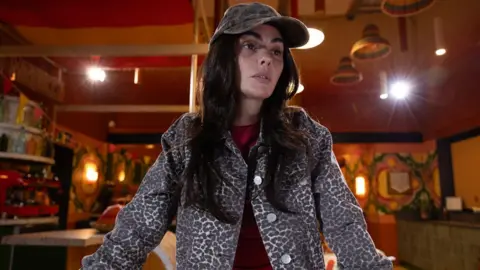 Lime Pictures/Channel 4 Jennifer Metcalfe as Mercedes McQueen, standing indoors with a baseball cap and looking forlorn