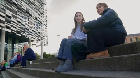 BBC/GEMMA LAISTER Two students sit on concrete steps. One has long brown hair and a light blue top and jeans. The second is wearing a black shiny jacket. In the background there are other students sitting down. 