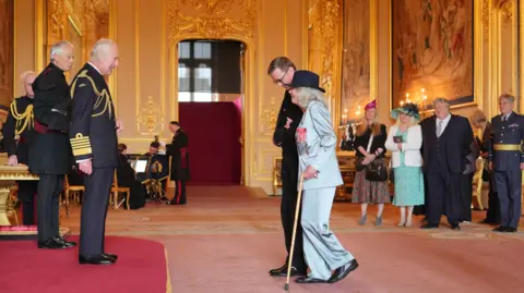 PA Dame Jilly Cooper, from Stroud, Author, is made a Dame Commander of the British Empire by King Charles III at Windsor Castle. She is walking towards the King, using a stick.