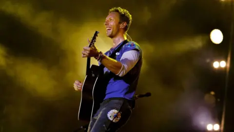 BBC/Steve Barney Chris Martin smiling and playing guitar wearing a blue top with black jeans with atmospheric lighting and mist behind him on stage