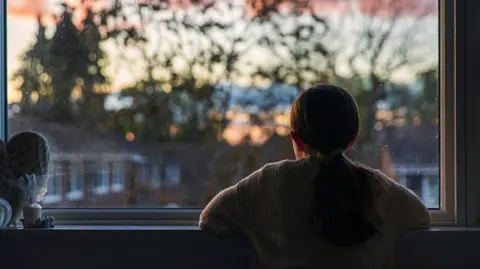 Getty Images A young girl looks out of a window at a darkening sky. Seen from behind, she is wearing a jumper and has her hair in a ponytail