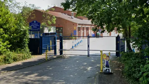 Google A street view image of St Monica's Catholic Primary School taken from outside its blue gates. The school building can be seen on the other side of the gates.