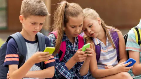 Getty Images Three children, each holding mobile phones, sitting beside each other on a wall. To the left is a boy with short brown hair, wearing a grey t-shirt and holding a yellow-backed phone. Two girls with shoulder length brown hair holding green and blue backed phoned are beside him.