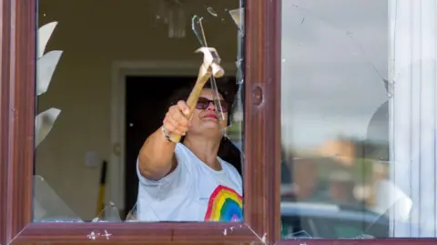 Getty Images A woman wearing a white T-shirt with a rainbow on the front stands inside a house. In front of her is a broken window pane. She is holding up a hammer and breaking the remaining bits of glass out of the pane.