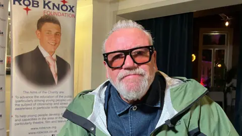 BBC / Jamie Moreland Actor Ray Winstone is wearing a denim shirt and green jacket with tinted glasses. Behind him is a poster that says "The  Rob Knox Foundation" and has a picture of a young man in a suit and pink tie.