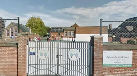 Google There is a brick wall and gates to Avenue Junior School in Norwich and behind is the school building with children playing on the playground.