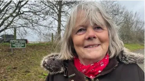 Andrew Turner/BBC Head and shoulders shot of Penny Carpenter smiling and wearing a brown winter coat with a fur trim and a red scarf. She is standing in a road with a green verge and trees behind.