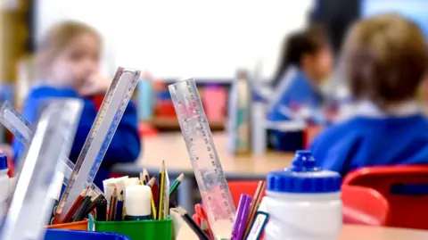 Getty Images Children sat in a classroom who are blurred out and can't be identified with rulers, pencils and super-glue in pots in the foreground.
