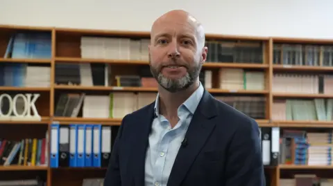 BBC/Brijesh Patel Tony Oulton looks at the camera smiling wearing a black suit jacket and light blue shirt. He has a grey and black short beard. Behind him there are shelves with rows of coloured books and folder. 