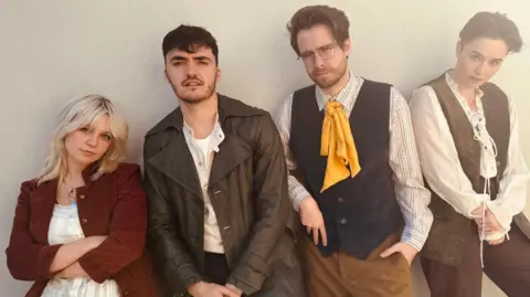 The John Godber Company Four people line up against a wall in old fashioned costumes as part of their roles in The Highwayman. From left, a blonde-haired woman has a red jacket on over a white top and has her arms folded; a man with short dark hair and stubble beard has a brown leather coat on over a white top; a man with brown hair and glasses is wearing a blue waistcoat with a yellow tie and has his left hand in his pocket; a woman with short dark hair has a brown waistcoat over a white top and is holding her hands together.