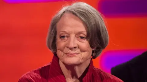 PA Media Dame Maggie Smith, a woman with dark grey bobbed hair, wearing a red shirt against a red and purple backdrop 