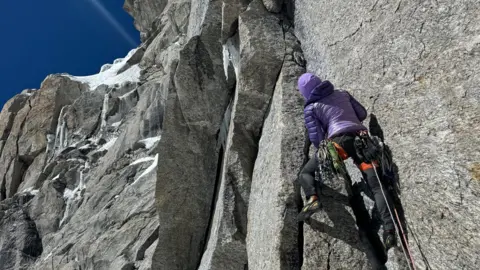 Fay Manners A woman dressed in purple with her back to the camera is climbing the side of a vast grey mountain with blue skies in the distance