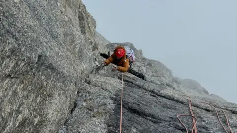 Fay Manners Taken from above, a woman in orange, her face obscured, is climbing the sheer face of a mountain, below her is thick fog obscuring the drop.