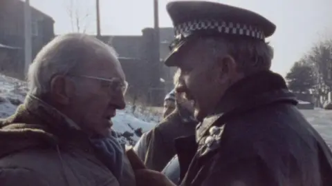 Kjell-Åke Andersson Man with grey hair and glasses to the left of frame is wearing a large coat and is facing off with a police officer. The officer is wearing his uniform which includes his black and white flat cap and a black overcoat. Around them are a few other miners who are standing on the picket line.