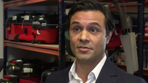 Dominic Ponniah wearing a white shirt and navy jacket standing in front of a number of Henry brand hoover vacuums