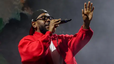 Getty Images Kendrick Lamar performing, singing into a mic while holding his left hand up towards the audience. He wears a red jacket, black-rimmed glasses and a black baseball cap twisted backwards. 