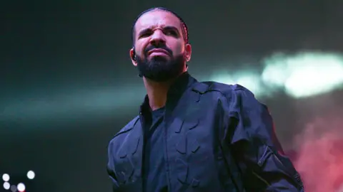 Getty Images Drake on stage. Drake wears his hair braided and has a short beard. He squints at the camera, his mouth tightly shut. He wears a black jacket over black T-shirt. 