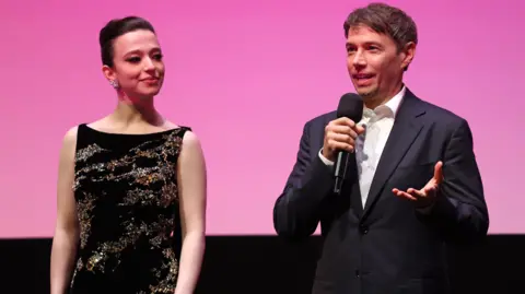 Getty Images Mikey Madison and Director Sean Baker on stage during the "Anora" Headline Gala during the 68th BFI London Film Festival at The Royal Festival Hall on October 11, 2024 in London, England. They are onstage introducing the film at the premiere. Baker is holding a microphone and talking to the audience. 