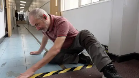 BBC/ Gemma Laister A man wearing a red polo shirt sticks yellow and black tape along the edge of a piece of carpet in a corridor at Patchway Community School.