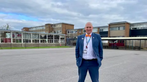 BBC/ Hazel Shearing Dave Baker, wearing a blue suit, white shirt and orange bow-tie, stands in the playground at Patchway School, in front of some of the buildings that will be demolished