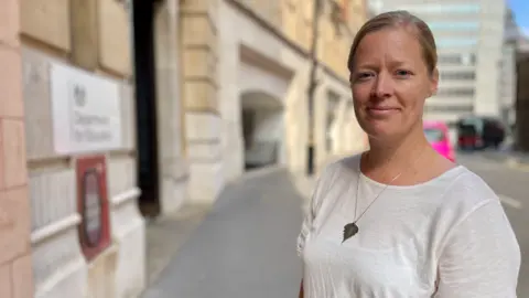 BBC/ Hazel Shearing Rebecca Larkin wears a white T-shirt with a silver necklace. It is a sunny day and she stands on the street outside the Department for Education in London.