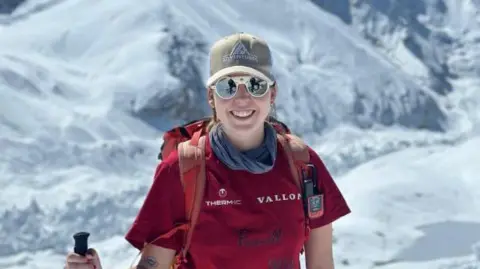 AGA Adventures Adriana Brownlee stands and smiles as she climbs a mountain, wearing a red t-shirt and backpack, brown hat and polarised sunglasses