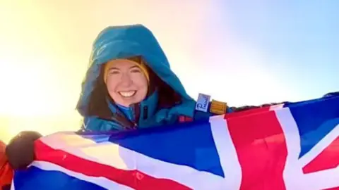 AGA Adventures Adriana Brownlee stands and smiles at the peak of a mountain in a blue snowsuit and yellow hat holding a Union Jack flag, with a sunrise behind her