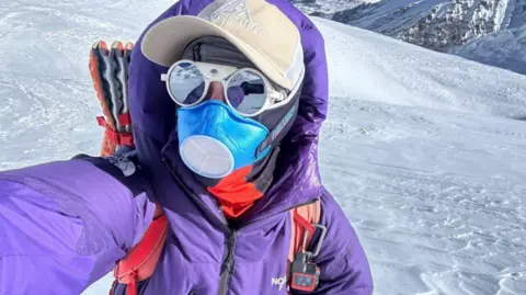 AGA Adventures Adriana Brownlee stands and looks towards the camera in a purple snowsuit and brown hat, while wearing a blue mask and polarised glasses, with snow behind her