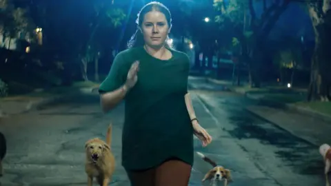 Searchlight Amy Adams in Nightbitch, seen running down the street surrounded by dogs