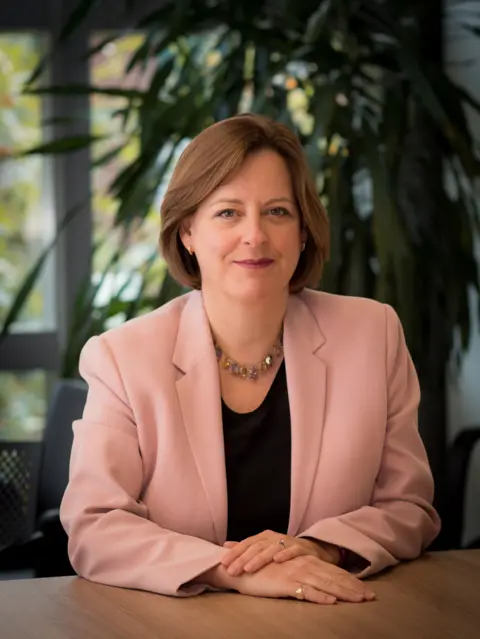 Ofcom / PA A woman in a salmon pink blazer sits at a table in a professional looking headshot style photograph