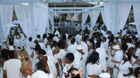 Getty Images Dozens of people dressed in all-white gather around a pool with diamond chandeliers and white sheer curtains at Sean "Diddy" Combs' White Party in 2007 