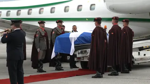 BBC News Alex Salmond coffin draped in Saltire surrounded by members of the North Macedonian military being loaded on to green and white plane.