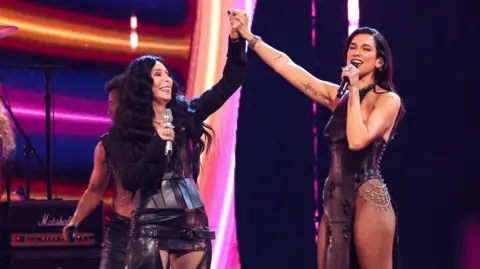 Getty Images Two women, Cher, and Dua Lipa. Cher has long black wavy hair and is wearing a short leather skirt with a buckle. She is holding the hand of the second woman, Dua Lipa. She is wearing a black sparkly swimming costume-type outfit. 