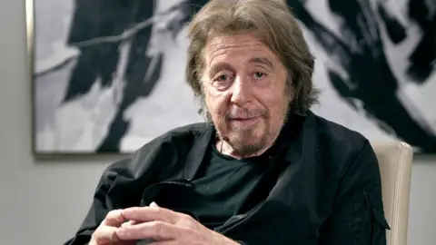 BBC Al Pacino sits and looks directly at the camera, his fingertips touching, with a piece of art behind him on the wall