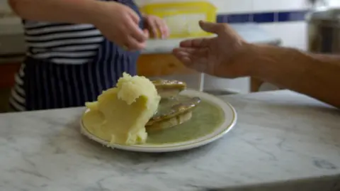 Phil Coomes/BBC Pie and mash with stacks of green liquor sauce on a white plate, being served in a cafe. The customer is about to receive some change from a man in a blue apron. Their faces are not visible.