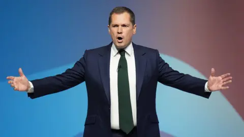 PA Media Robert Jenrick wearing a blue suit, delivering a speech on stage at the Conservative Party conference in Birmingham