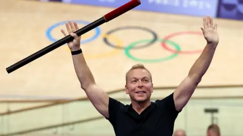 Getty Images Chris Hoy, wearing a black t-shirt, holds a baton in the air during a ceremony at a velodrome in Paris. Behind him is the velodrome with the five Olympic rings