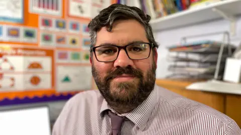 BBC Headteacher in a classroom wearing glasses and a striped shirt. The background display features mathematics.