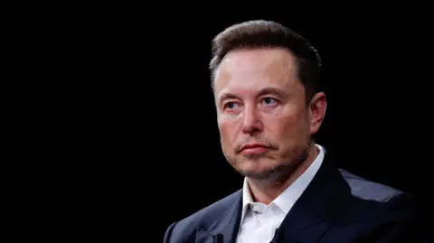 Reuters Elon Musk, wearing a black suit jacket and white shirt, sits against a black backdrop on stage at an event.