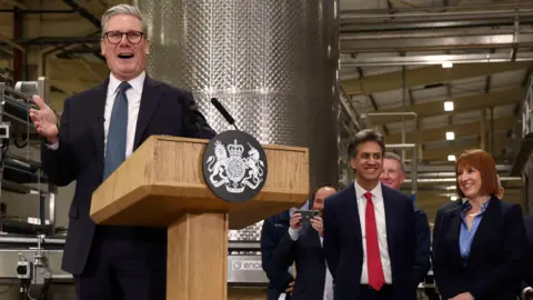 Darren Staples/PA Sir Keir Starmer makes a speech in front of a wooden podium. He has his hand clenched. He is wearing a suit and navy tie. Rachel Reeves, in a blue shirt and suit jacket, and Ed Miliband, in a red tie and white shirt and navy suit, look on during a tour of a factory in Cheshire.