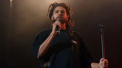 Getty Images J Cole on stage. J Cole holds has dreads and a short beard. In his right hand, he holds a mic to his face, holding the mic stand with his left. 