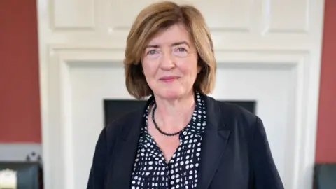 PA Media Sue Gray wearing a navy blouse with white spots, a small, beaded navy necklace, and a navy jacket smiles slightly in a Downing Street office.