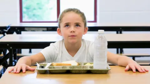 Getty Images Stock image of a young girl with light hair, wearing a white top. She looks frustrated with hands on a school table in front of a tray of food and bottle of water.