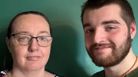 BBC A women with dark purple glasses stands next to a man with a brown beard and brown necklace