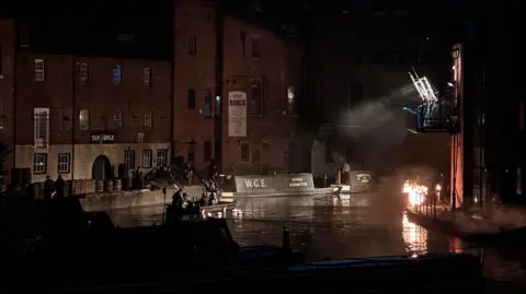 Dyson Media A canal at night with redbrick warehouse-style buildings on either side. There are large professional studio lights installed on a balcony and pointing at a group standing on a towpath. A fire has also been created on one side of the canal.