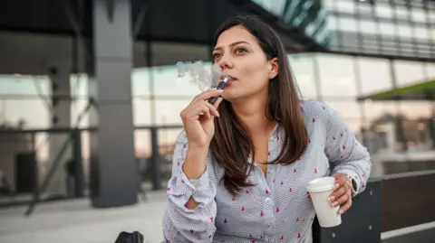 Getty Images Young woman with brown hair vaping. She has a coffee cup in one hand and is sitting outside a glass-fronted building. 