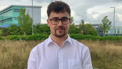 BBC Alex Whinfrey-Gibson stares at the camera. He has brown hair and a brown beard and is wearing a white, open-necked shirt and dark framed glasses