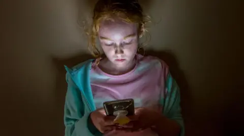 Getty Images A young long haired redhead girl with face lit up looking into mobile phone screen in a dark room, against a flat wall, with sad facial expression