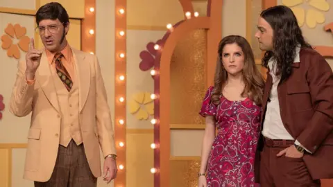 Netflix Tony Hale in a beige and brown 70s suit, Anna Kendrick in a red paisley dress and Daniel Zovatto in a brown 70s suit in Netflix's Woman of the Hour